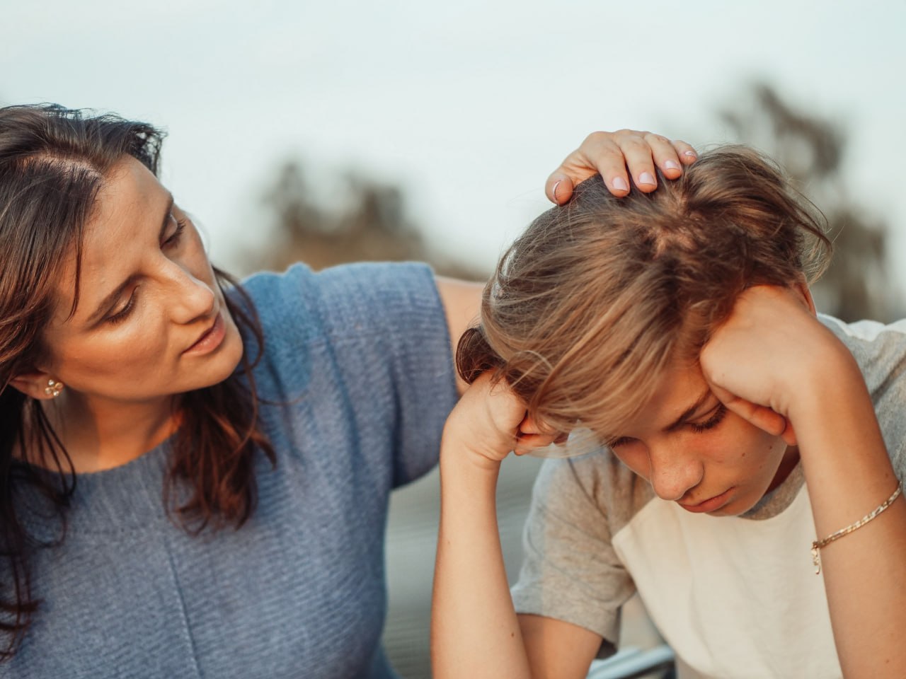 A mother resting her hand on her teenage son's lowered head as they sit outside together